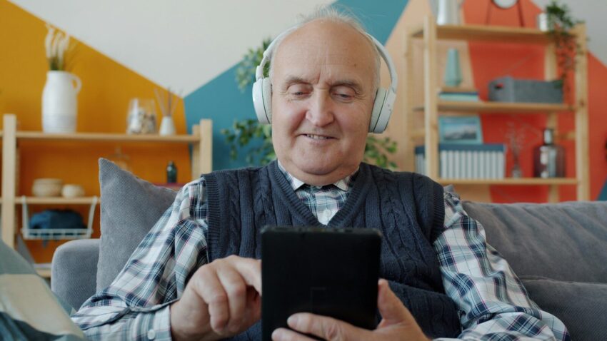 Elderly man with headphones uses a tablet on couch